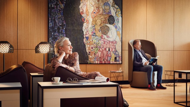 A blonde woman is sitting in the Vienna Lounge in front of a Klimt painting.