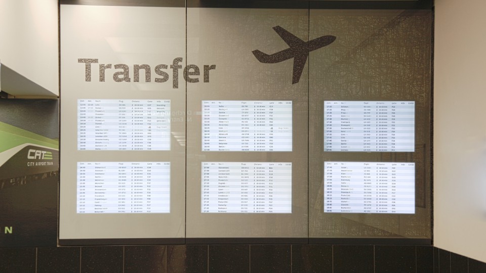 Eine digitale Anzeigetafel in einem Flughafenbereich mit dem Schriftzug "Transfer" und einem stilisierten Flugzeugsymbol darüber. Die Tafel zeigt mehrere Bildschirme, die Ankunfts- und Abflugdaten für verschiedene Flüge listen. Links unten ist ein Teil eines Logos zu sehen, das "CAT City Airport Train" anzeigt.