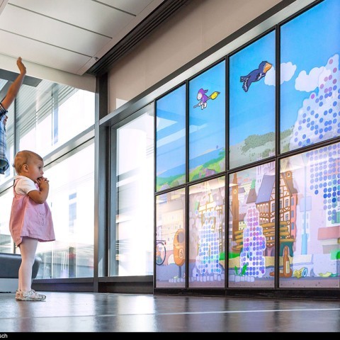 Zwei Kinder stehen vor einer großen interaktiven Wand mit bunten Animationen. Die Wand zeigt eine Szene mit animierten Vögeln, die am Himmel fliegen, und eine Stadtansicht im Hintergrund. Das jüngere Kind steht näher an der Wand und beobachtet konzentriert, während das ältere Kind im Hintergrund die Arme hebt. Die Umgebung sieht modern aus, möglicherweise ein öffentlicher Bereich oder ein Flughafen.