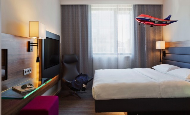 Modern eingerichtetes Hotelzimmer im Moxy Hotel mit einem großen Doppelbett, grauen Wandpaneelen und einem großen Fenster mit blickdichten Vorhängen. Neben dem Bett steht eine stylische Lampe auf einem kleinen Nachttisch. Gegenüber dem Bett befindet sich ein Flachbildfernseher an der Wand und ein bequemer Sessel. Auffällig ist ein rotes Modellflugzeug, das scheinbar im Flug im Raum schwebt.