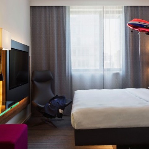 Modernly furnished hotel room at Moxy Hotel featuring a large double bed, gray wall panels, and a large window with opaque curtains. There is a stylish lamp on a small bedside table next to the bed. Opposite the bed, there is a flat-screen TV mounted on the wall and a comfortable armchair. A striking feature is a red model airplane that appears to be flying through the room.