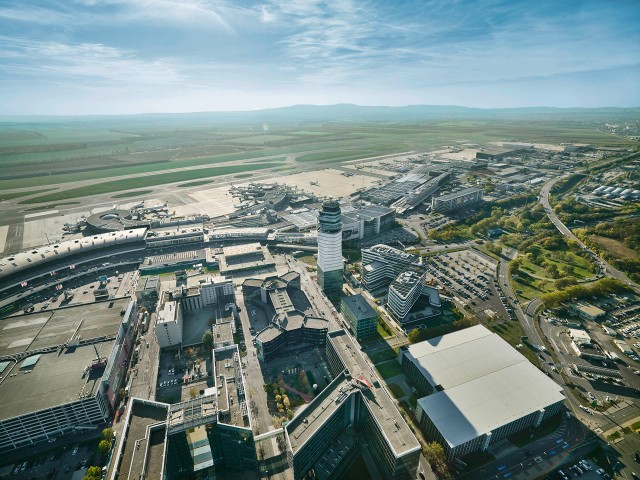 Flughafen Wien Luftaufnahme: Luftaufnahme des Flughafen Wien an einem klaren Tag. Im Zentrum befindet sich der markante Tower, umgeben von Terminalgebäuden, Flugsteigen und startbereiten Flugzeugen. Im Hintergrund sind grüne Felder, Landebahnen und die hügelige Landschaft Niederösterreichs zu sehen. Rechts im Bild verlaufen Zufahrtsstraßen und Parkplätze, links moderne Büro- und Hotelgebäude.
